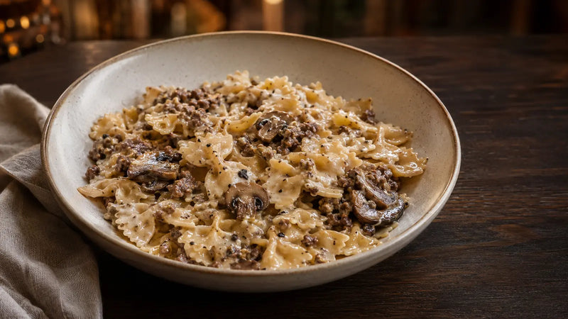 Un bol de pâtes crémeuses avec de la viande hachée et des champignons, servi sur une table en bois foncé avec une serviette pliée.