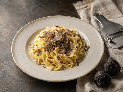 Tagliatelles avec des copeaux de truffe noire, des truffes entières et une mandoline à truffes sur un plan de travail en pierre.